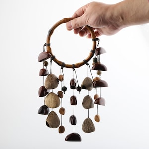 May include: A handcrafted wind chime featuring a circular bamboo frame. Various shapes and sizes of brown, beige, and dark brown wooden beads and shells are suspended from the frame with black string. The wind chime is held by a hand.