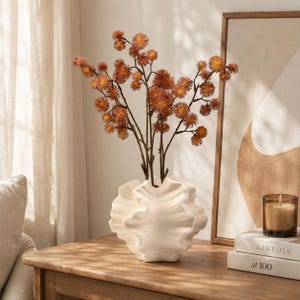Pode incluir: Um vaso de cerâmica branca com uma textura ondulada semelhante a uma concha contém vários ramos finos castanho-escuros adornados com pequenas flores redondas de cor laranja acastanhada. O vaso tem uma borda larga e ondulada e base arredondada.