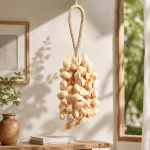 May include: A hanging seashell decoration. The decorative piece is made of numerous beige seashells, suspended from a woven rope loop. The shells are arranged in a cascading cluster, creating a natural, coastal aesthetic. The background includes a window and a wooden frame.