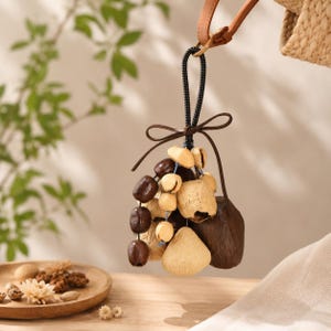 May include: A decorative charm featuring a cluster of natural elements. The charm includes dark brown seed pods, light brown seed pods, and beige seed pods, tied together with a brown leather bow and black cord. The charm is suspended from a leather strap.
