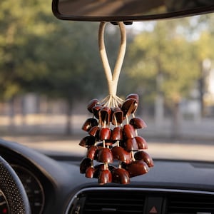 May include: A car air freshener with a cluster of dark brown, acorn-shaped beads hanging from a cream-colored rope. The air freshener is suspended from a car's rearview mirror, with the dashboard and steering wheel visible.