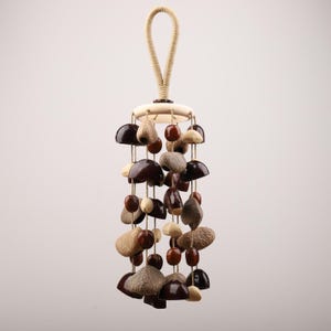 Könnte beinhalten: Hängemobile mit Holzring und Schlaufe. Es zeigt eine Sammlung natürlicher Elemente, darunter dunkelbraune, beige und cremefarbene Samenkapseln und Perlen, die an Schnüren befestigt sind. Das Design ist erdig und organisch, geeignet für die Inneneinrichtung.