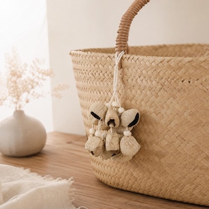 May include: A woven straw tote bag with a rounded handle and a decorative tassel. The bag is a natural beige color and has a textured pattern. The tassel features beige bell-shaped ornaments and white beads.