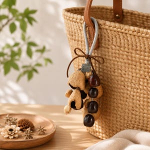 May include: A woven straw tote bag with a brown leather handle and a decorative charm. The charm features a cluster of dried seed pods and dark brown, round beads, tied with a brown leather cord.