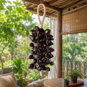 May include: A dark brown wind chime made of small, rounded wooden pieces suspended from a natural rope. The chime hangs from a woven ceiling, with greenery and a table in the background.
