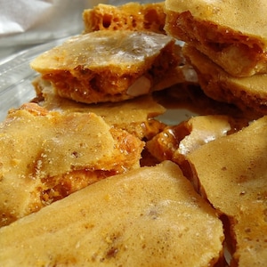 May include: Close-up of a pile of golden-brown honeycomb candy. The candy has a bubbly, porous texture and a sweet, caramelized appearance. The pieces are irregularly shaped and appear crispy.