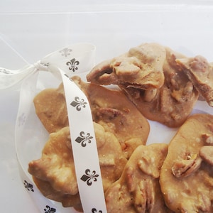 May include: A clear plastic container filled with pecan pralines. The light brown candies are tied with a white ribbon printed with black fleur-de-lis symbols. The pralines have a glossy finish.