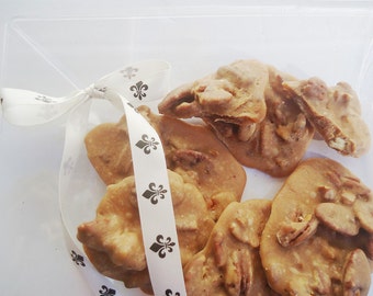 May include: A clear plastic container filled with pecan pralines. The light brown candies are tied with a white ribbon printed with black fleur-de-lis symbols. The pralines have a glossy finish.