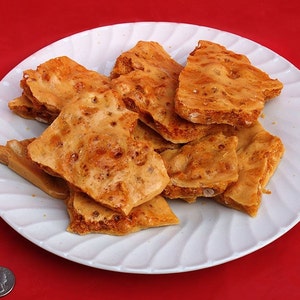 May include: A white plate piled with pieces of golden-brown honeycomb candy. The candy has a bubbly texture and a light, airy appearance. The plate sits on a bright red surface, with a quarter coin for scale.