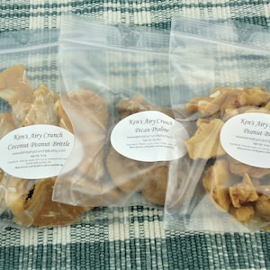May include: Three clear plastic bags filled with Ken's Airy Crunch brittle. The bags are labeled with the product names: Coconut Peanut Brittle, Pecan Praline, and Peanut Brittle. The brittle pieces are light brown and appear crunchy.