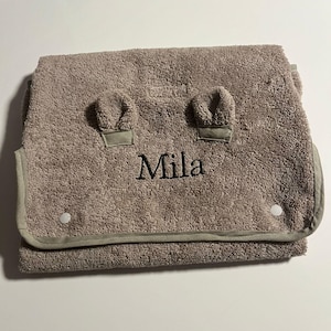 Peut inclure: Serviette à capuche pour bébé taupe avec le nom "Mila" brodé en noir. La serviette présente deux petits accents en forme d'oreilles et une bordure gris clair. Elle se ferme avec deux boutons-pression blancs et est en matière douce et absorbante.