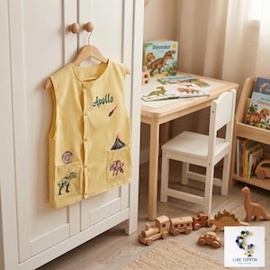 Peut inclure: Un gilet sans manches jaune à petit motif à carreaux, orné du nom brodé Apollo, d'un volcan et de diverses illustrations de dinosaures. Le gilet présente une fermeture boutonnée sur le devant et deux poches plaquées sur la partie inférieure.