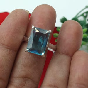 May include: A silver ring featuring a large, rectangular, teal-colored gemstone. The gemstone is set with four prongs. The ring is held up by a person's fingers, with a red and green background.
