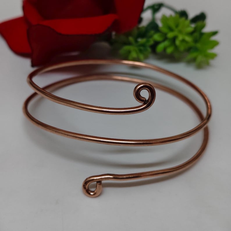 May include: Copper-coloured arm cuff bracelet with a spiral design. The bracelet wraps around the arm with a looped end and a decorative swirl. The background includes a red rose and green foliage.