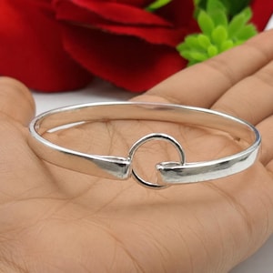 May include: A polished silver bangle bracelet featuring a smooth, continuous band that terminates in a circular loop design. The metal has a reflective, high-shine finish and a rounded profile, creating a clean and simple aesthetic for the wrist.