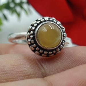 May include: A silver ring featuring a round, light yellow gemstone. The gemstone is set in a bezel surrounded by a textured, beaded design. The ring's band is smooth and simple, creating a contrast with the ornate setting.