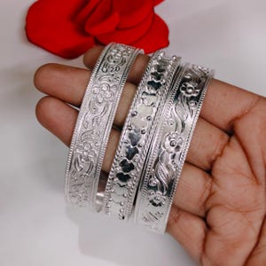 May include: Four silver bangles with detailed floral and leaf patterns are shown. The bangles are held in a hand, with a red rose in the background. The bangles feature intricate designs, showcasing a classic jewellery style.