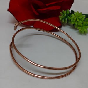 May include: A copper-coloured, minimalist bracelet with a spiral design. The bracelet is made of a single, continuous piece of metal that wraps around the wrist twice. A red rose and green floral accents are in the background.