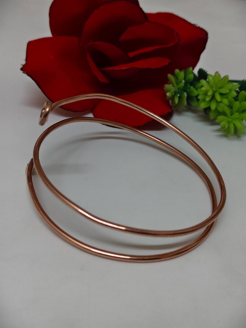 May include: A copper-coloured, minimalist bracelet with a spiral design. The bracelet is made of a single, continuous piece of metal that wraps around the wrist twice. A red rose and green floral accents are in the background.