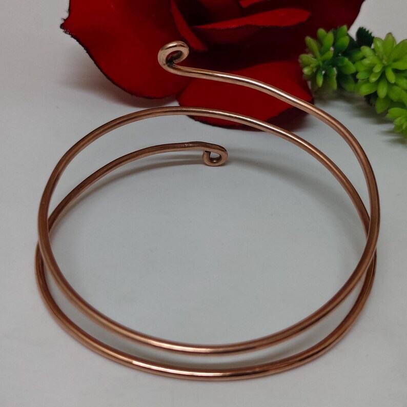 May include: A copper-coloured arm cuff bracelet with a spiral design. The bracelet wraps around the arm with two parallel bands and a decorative spiral at the top. The jewellery is displayed with a red rose and green foliage.