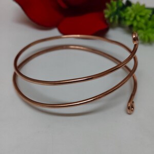 May include: A copper-coloured spiral arm cuff bracelet. The bracelet is made of a single, continuous piece of metal that coils around the arm. The ends of the bracelet are rounded. The bracelet is displayed on a white surface.