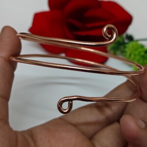 May include: A copper-coloured arm cuff with a spiral design. The cuff wraps around the arm with two curved ends, one at the top and one at the bottom. The background includes a red rose and green foliage.