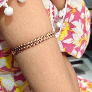 May include: A copper-colored bracelet with a chain-link design. The bracelet is worn on an arm, and the links are closely connected. The bracelet is a warm, metallic color and appears to be made of metal.