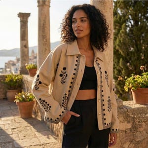 Puede incluir: Chaqueta beige con bordados florales negros y un diseño de borde geométrico. La chaqueta tiene cuello y cierre de botones. Las mangas y los paneles frontales presentan el estampado floral. La persona de la imagen lleva un top negro y pantalones negros.