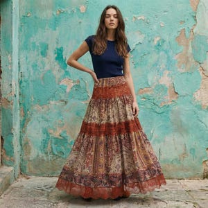 Può includere: Una donna indossa un top blu navy a maniche corte e una gonna lunga a balze. La gonna presenta un motivo paisley nei toni del marrone, arancione e viola, con finiture in pizzo sull'orlo. Lo sfondo è un muro turchese.