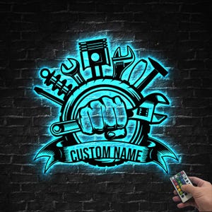 Peut inclure: Enseigne au néon noire et bleue représentant des outils de mécanicien. Le design comprend un piston, des clés et un poing tenant une clé, avec le texte "CUSTOM NAME" sur une bannière. L'enseigne est fixée sur un mur de briques sombres.