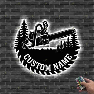 Peut inclure: Un design de tronçonneuse noire avec les mots "CUSTOM NAME" en blanc, entouré d'une silhouette de forêt. Le design est éclairé d'une lueur blanche, sur fond de mur de briques. Une télécommande est visible.