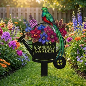 Könnte beinhalten: Ein schwarzer Metall-Gartenstecker mit einem bunten Vogel, der auf einer Gießkanne mit Blumen sitzt. Der Text "GRANDMA'S GARDEN" ist auf der Gießkanne zu sehen. Der Vogel hat grünes, rotes und blaues Gefieder. Der Stecker steht in einem Blumengarten.