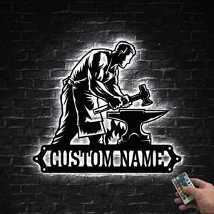 May include: Black and white metal sign depicting a blacksmith at work, hammering on an anvil. The sign includes the text "CUSTOM NAME" and is mounted on a brick wall. A remote control with color options is visible.