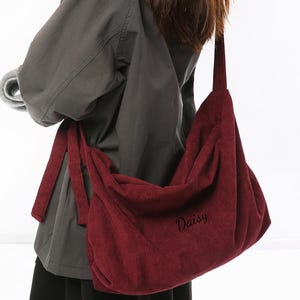 May include: A burgundy corduroy shoulder bag with the word "Daisy" embroidered on the front. The bag has a long strap and is worn over the shoulder. The bag is a deep red color and has a soft, textured appearance.