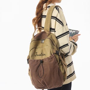Puede incluir: Mochila de lona en color beige y marrón, con el nombre "Justin" impreso en la parte delantera. La mochila tiene correas ajustables, un asa superior y un bolsillo con cordón. Ideal para llevar libros y material escolar.