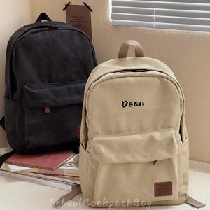 以下が含まれることがあります： 2つのキャンバスバックパック。1つは黒、もう1つはベージュで、前面に「Dean」という名前が刺繍されています。どちらもフロントポケット、上部のハンドル、調節可能なストラップが付いています。ベージュのバックパックの下部には小さな茶色のパッチがあります。「SchoolBackpackBag」というテキストが下にあります。