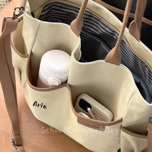 Könnte beinhalten: Beige Canvas-Tragetasche mit braunen Verzierungen und Riemen. Die Tasche hat mehrere Taschen, darunter eine mit einer weißen Wasserflasche und eine weitere mit einem Telefon. Das Wort "Aria" ist auf der Vorderseite geschrieben. Der Text "School Backpack Blog" ist ebenfalls sichtbar.