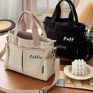 以下が含まれることがあります： 2つのキャンバス製トートバッグ。1つはベージュで「Callie」という名前と「School Backpack Bag」という文字が入っています。もう1つは黒で「Puff」という名前が入っています。どちらのバッグにもハンドル、ショルダーストラップ、複数のポケットが付いています。