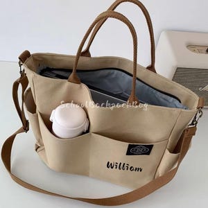 May include: Beige canvas tote bag with brown handles and a shoulder strap. Features multiple exterior pockets, one holding a white water bottle. The name "William" is embroidered in black on the front. The bag is open, revealing a dark interior.