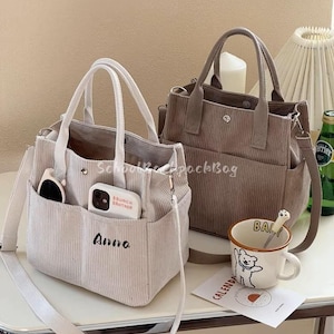 May include: Two corduroy tote bags in beige and taupe, each with a top handle and a detachable shoulder strap. The beige bag has the name "Anna" embroidered on the front. A mug with a bear graphic and a spoon rests on a table.