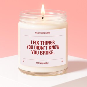 Könnte beinhalten: Eine brennende Sojawachskerze in einem durchsichtigen Glas. Die Kerze hat ein weißes Etikett mit dem Text "I FIX THINGS YOU DIDN'T KNOW YOU BROKE." Die Kerze ist 255g und hat eine Brenndauer von 60 Stunden.