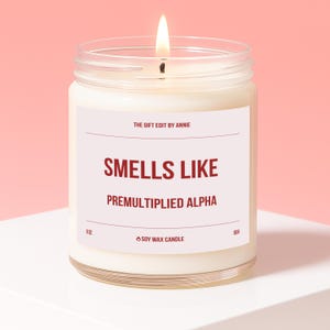 May include: A lit soy wax candle in a clear glass jar. The label reads "Smells Like Premultiplied Alpha" with the text "The Gift Edit by Annie" above. The candle is 9 oz and has a burn time of 60 hours. The background is pink.