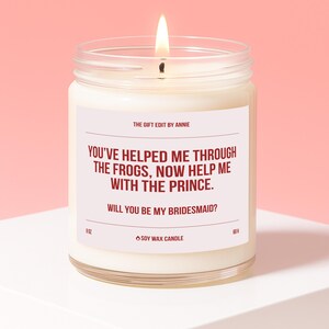May include: A lit soy wax candle in a clear glass jar. The candle has a white label with the text "YOU'VE HELPED ME THROUGH THE FROGS, NOW HELP ME WITH THE PRINCE. WILL YOU BE MY BRIDESMAID?" The candle is 9 oz.