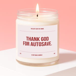 May include: A lit soy wax candle in a clear glass jar. The label reads "THANK GOD FOR AUTOSAVE." and "THE GIFT EDIT BY ANNIE." The candle is 9 oz and has a 60-hour burn time. The background is a soft pink.