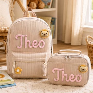 May include: A beige corduroy backpack and a matching rectangular lunch bag, both featuring the name Theo in pink yarn lettering and a small plush lion face appliqué. The backpack has a yellow flower accent on the front pocket zipper pull.
