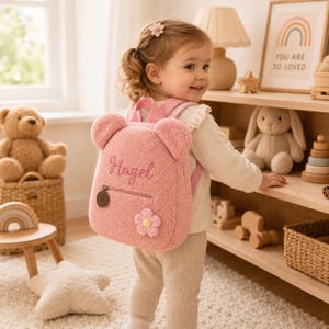 Könnte beinhalten: Ein weicher, rosafarbener Rucksack aus Plüsch mit Bärenohren, auf dessen Vordertasche der Name Hazel in Schreibschrift gestickt ist. Der Rucksack ist mit einer kleinen gehäkelten Blume mit gelber Mitte und einem runden braunen Reißverschluss versehen.