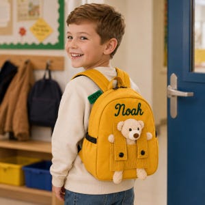 May include: A mustard yellow backpack featuring the name Noah embroidered in dark green script. The bag has a front pocket with a plush beige teddy bear attached, and a small green tag on the shoulder strap with the word HAPPY printed on it.