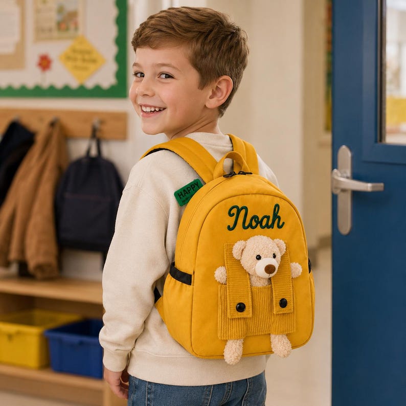 May include: A mustard yellow backpack featuring the name Noah embroidered in dark green script. The bag has a front pocket with a plush beige teddy bear attached, and a small green tag on the shoulder strap with the word HAPPY printed on it.
