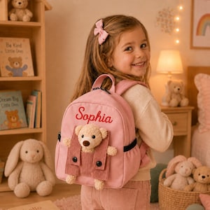 Könnte beinhalten: Ein hellrosa Rucksack aus Segeltuch mit dem in roter Schreibschrift gestickten Namen Sophia. Die Vorderseite der Tasche hat eine kleine Tasche mit einem befestigten Plüschbären, der eine Cord-Latzhose mit zwei schwarzen Knöpfen trägt.