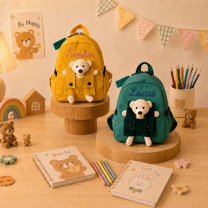 May include: A yellow backpack with the name Sophie and a green backpack with the name Lucas, each featuring a small plush bear in a front pocket. Both bags have top handles, black zippers, and side pockets, displayed on wooden stands.
